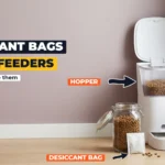 White automatic pet feeder with its lid open beside a glass kibble jar and a small white desiccant packet on the floor, with a tabby cat looking on