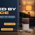 White smart pet feeder with a small smart speaker and a phone beside it on a wooden floor and a Labrador-type dog looking up at it, against a navy blue wall