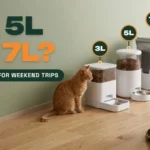 Three white automatic pet feeders of different sizes lined up on a wooden floor with a ginger cat looking at the smallest one and a travel duffel bag nearby, against a sage green wall